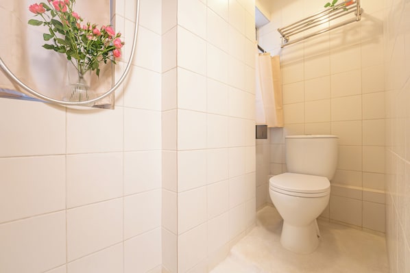 A bathroom with white tiles, a toilet, a towel rack, and a mirror with a vase of flowers. - Apple Inn (Causeway Bay) (Hong Kong)