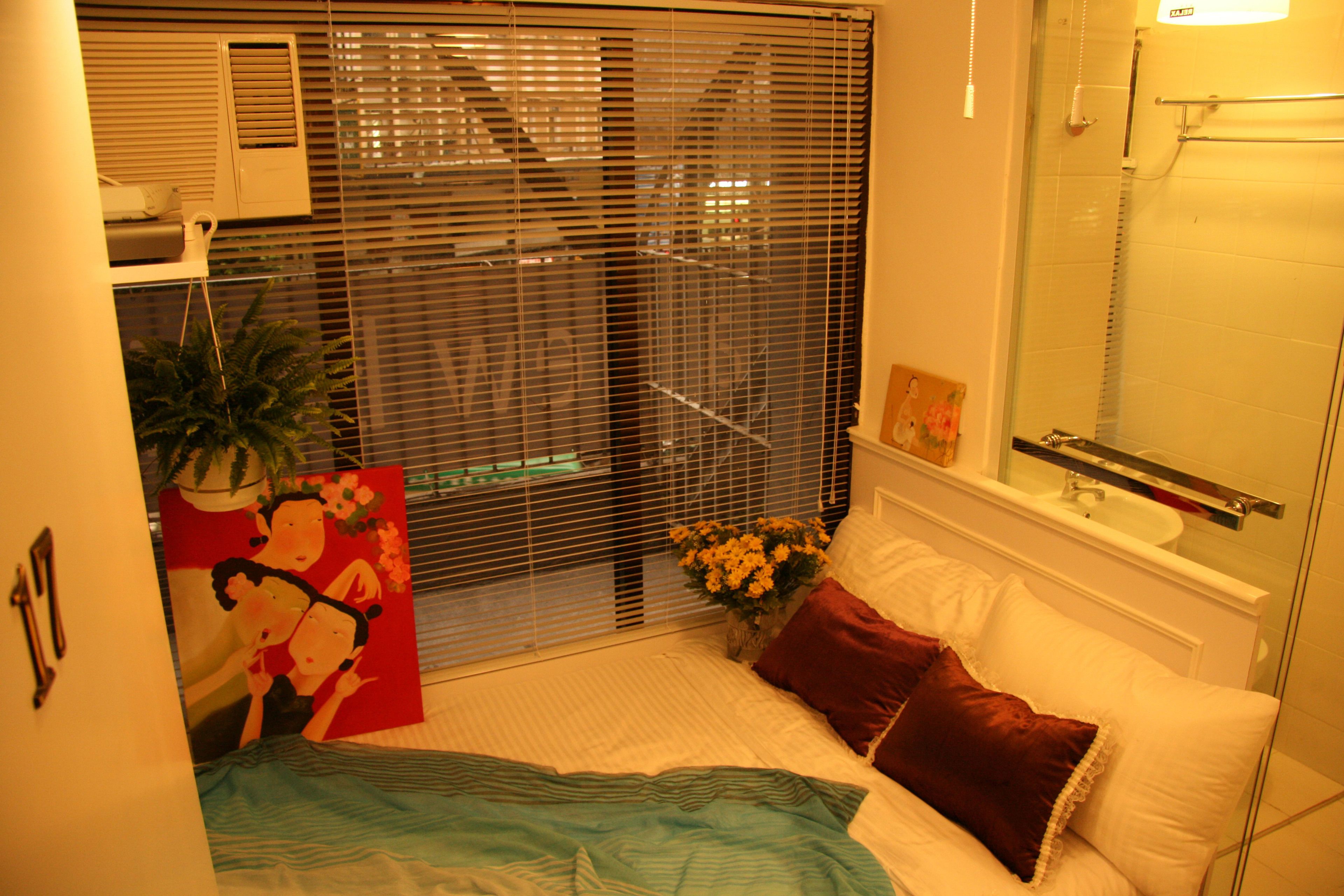 A bedroom with a bed, a window with blinds, a bedside table, and a bathroom with a shower.