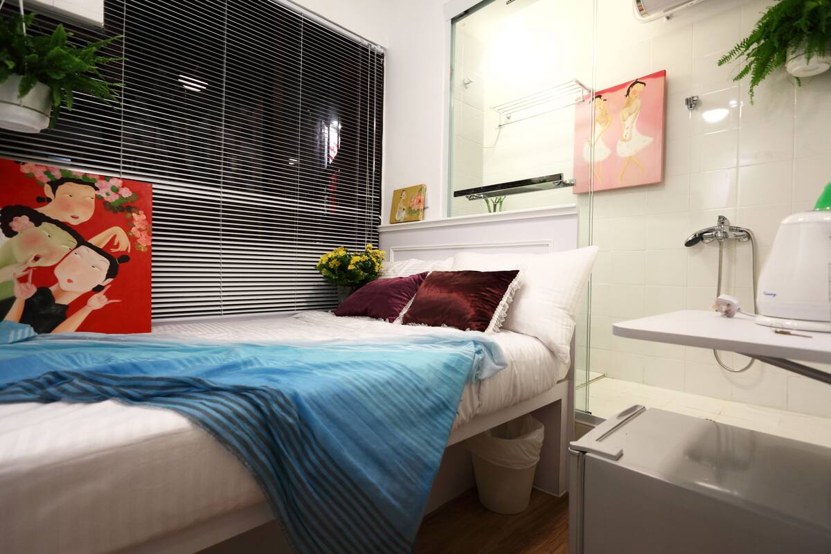 A bedroom with a bed, a window with blinds, a bedside table, a mirror, and a small plant.