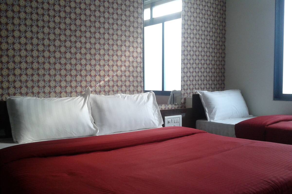 A hotel room with two beds, each with red bedding and white pillows. The room has patterned wallpaper and a window with curtains.