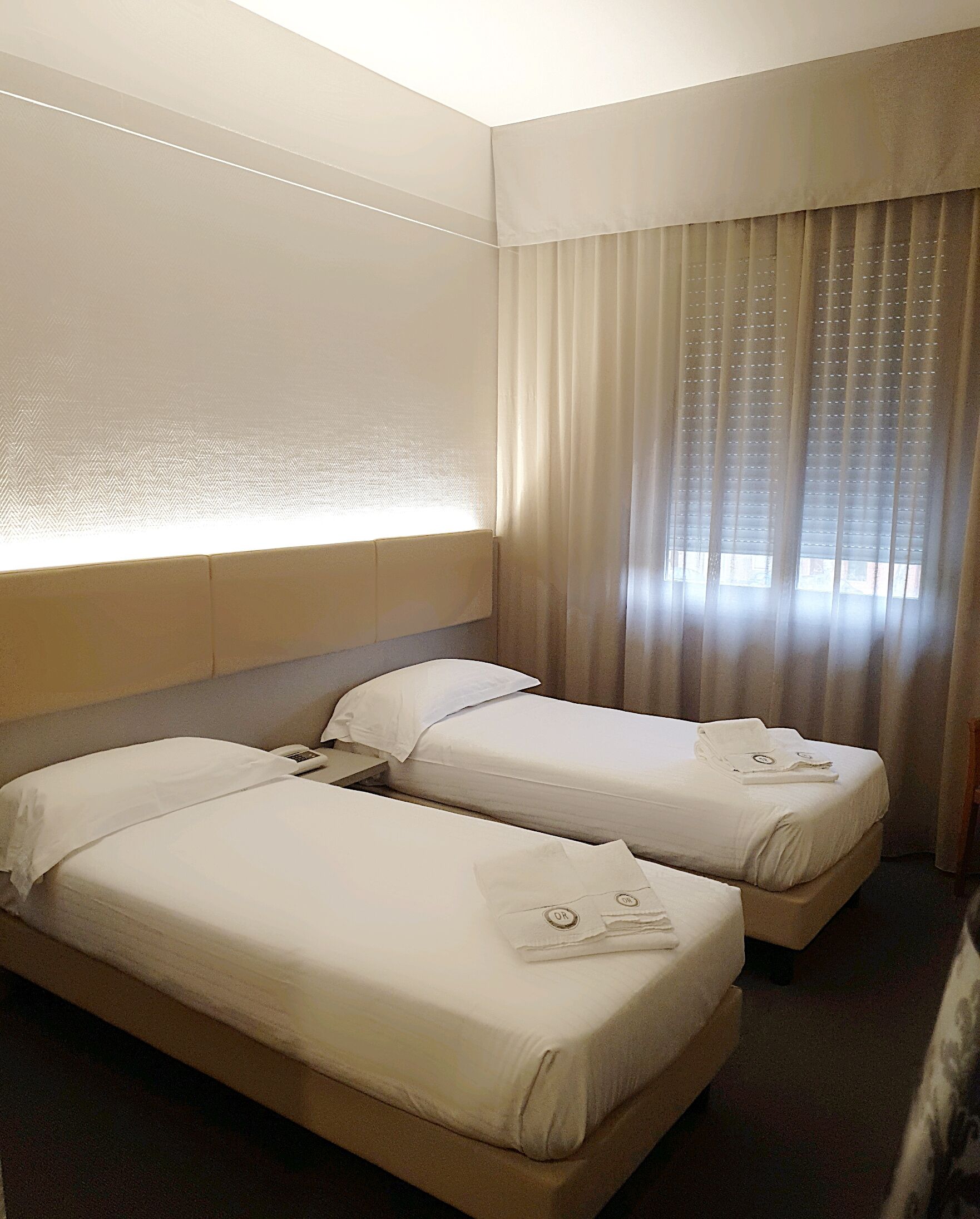 A hotel room with two single beds, a headboard, a bedside table with a phone, and a window with curtains.