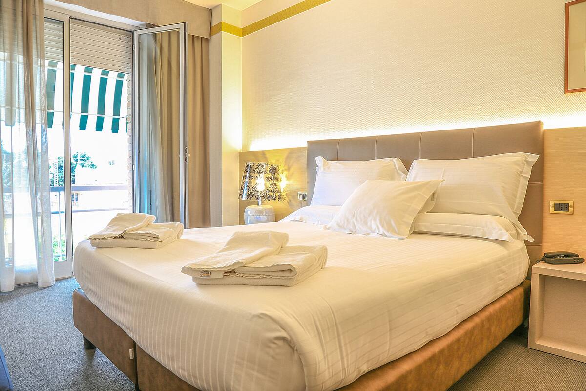 A neatly made bed with pillows and folded towels, a bedside table with a lamp, and a view through a sliding door to the outside.