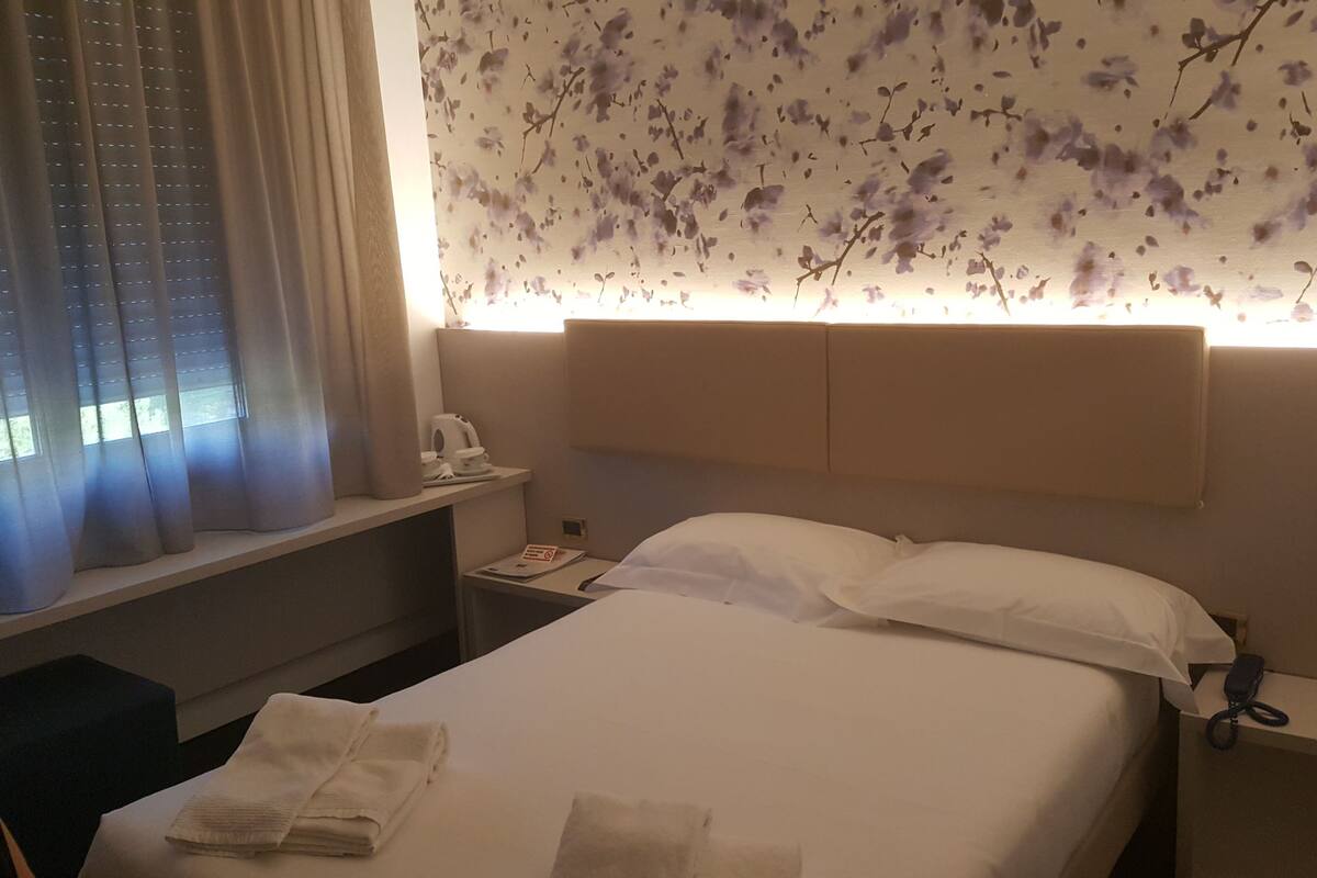 A hotel room with a bed, a nightstand, a window with curtains, and a wall with a floral pattern.
