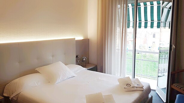 A hotel room with a large bed, a nightstand, a window with curtains, and a view of buildings outside. - Hotel Augustus (Fano)