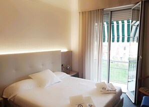 A hotel room with a large bed, a nightstand, a window with curtains, and a view of buildings outside. - Hotel Augustus (Fano)