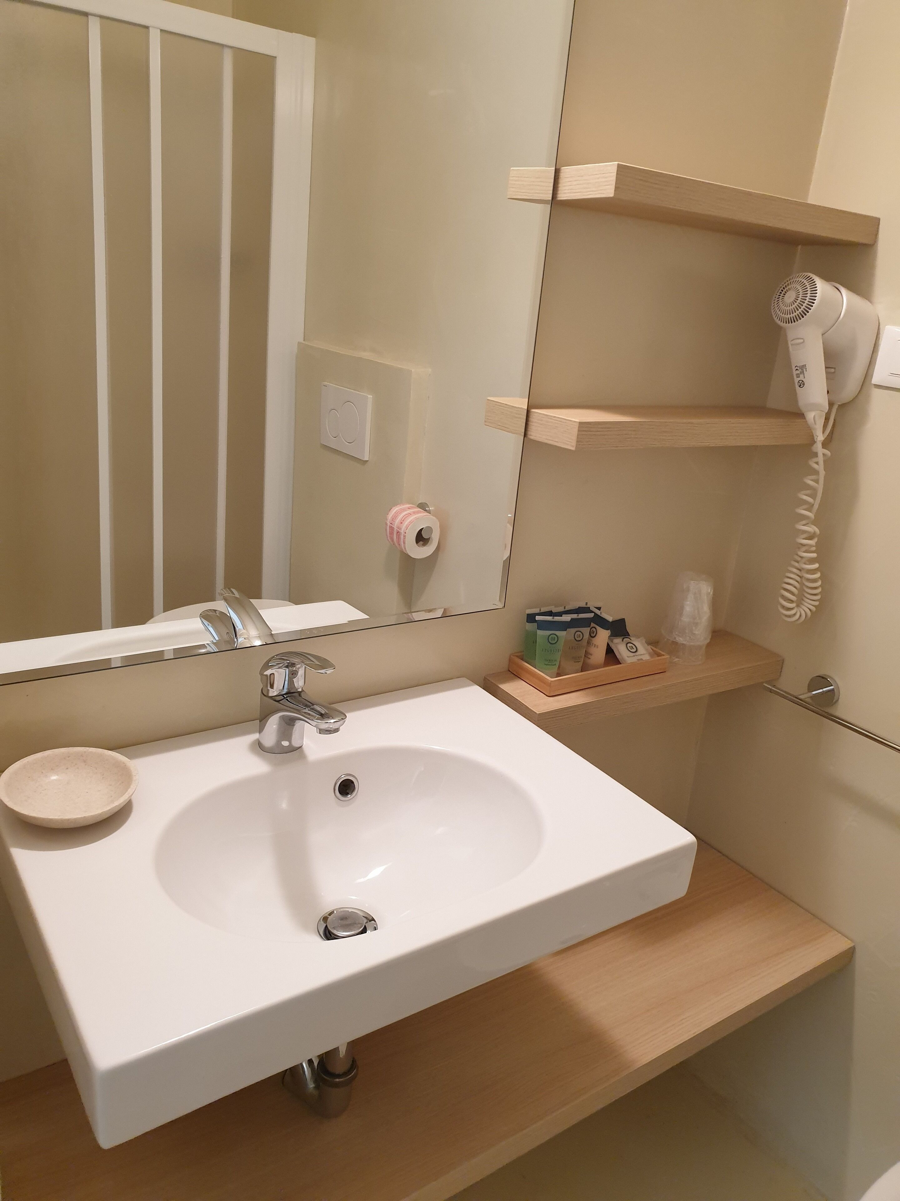 A bathroom with a white sink, a mirror, a towel dispenser, and a hairdryer on a wooden shelf.