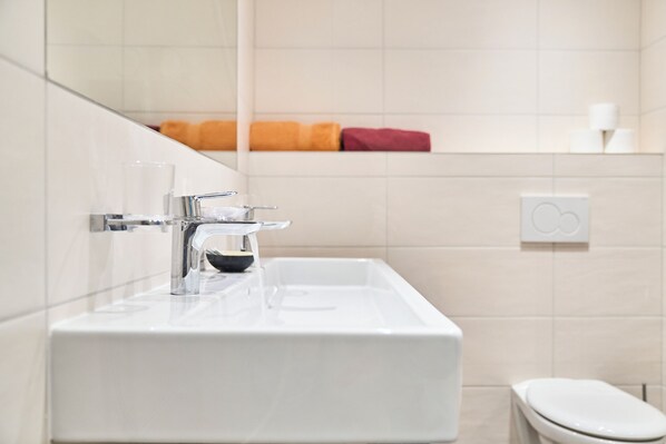 Une salle de bain avec un lavabo blanc, un miroir et des toilettes. On aperçoit des serviettes au fond.
