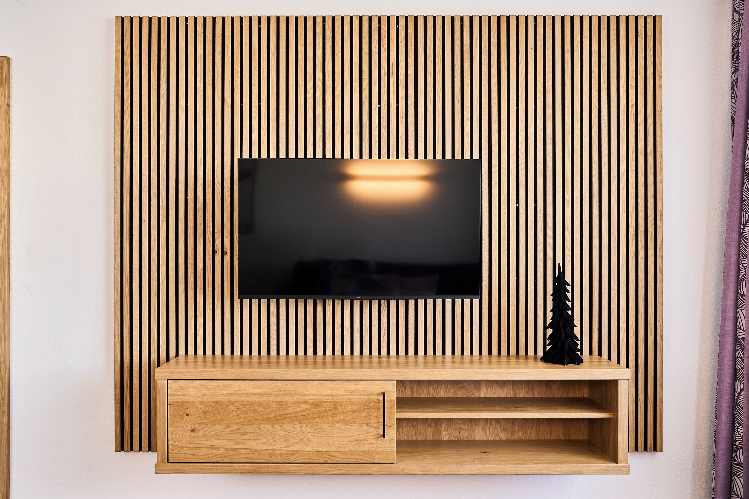 Un meuble de divertissement moderne, avec un téléviseur à écran plat fixé sur un mur composé de lattes de bois, un meuble en bois en dessous et un petit sapin de Noël noir.