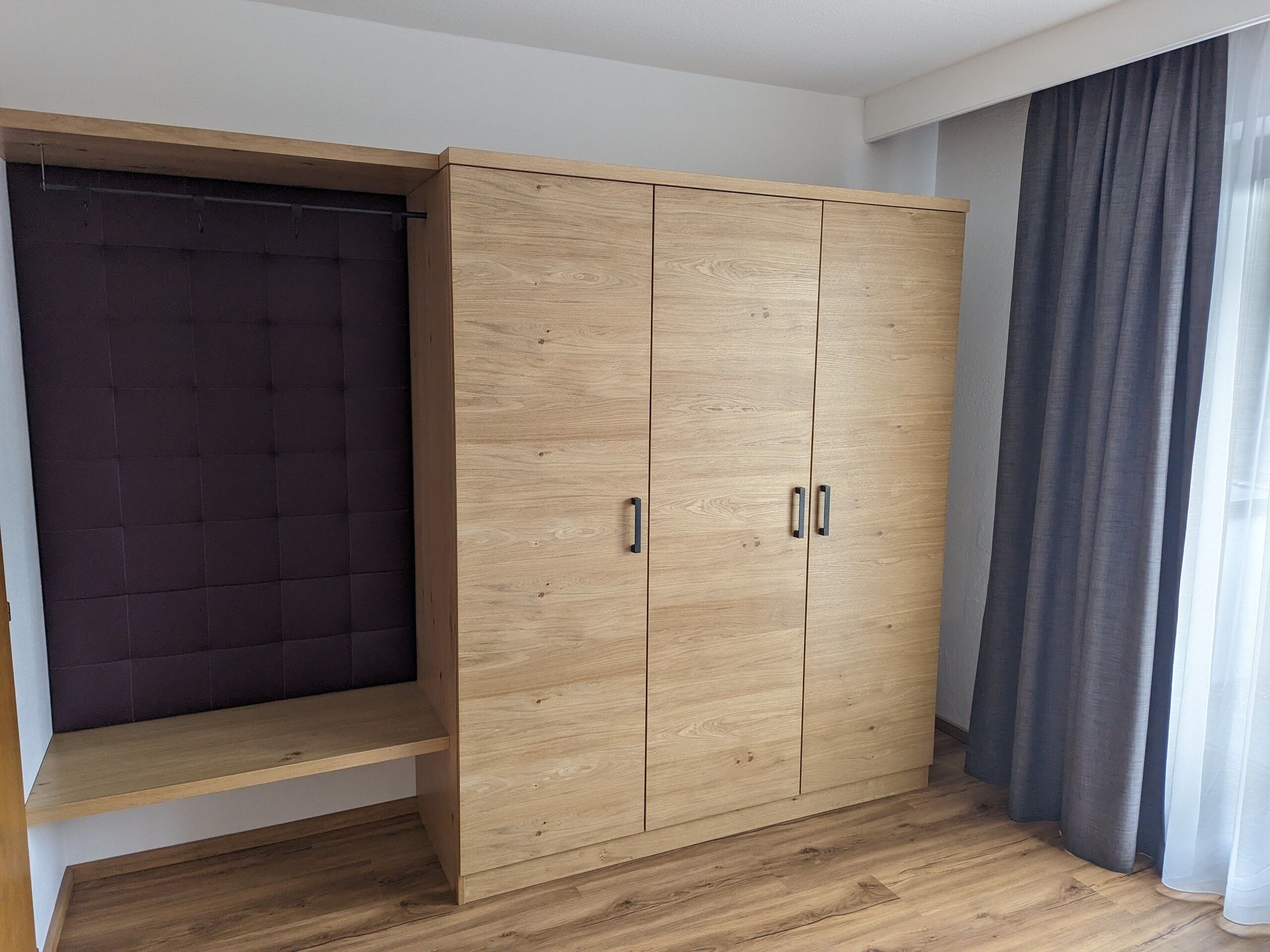 A room with a wooden wardrobe, a bench, and a window with curtains.