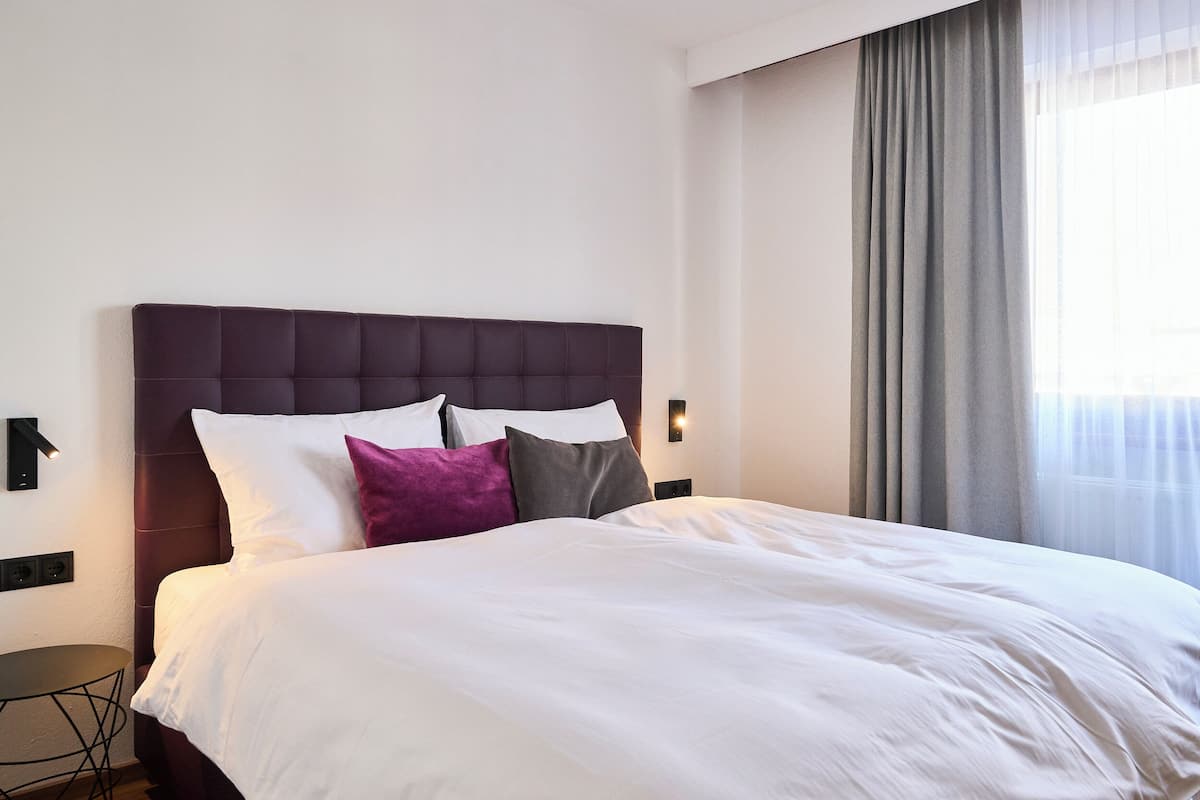 Ein ordentlich bezogenes Bett mit weißer und grauer Bettwäsche, einem dunklen Kopfteil und einem violetten Kissen.
