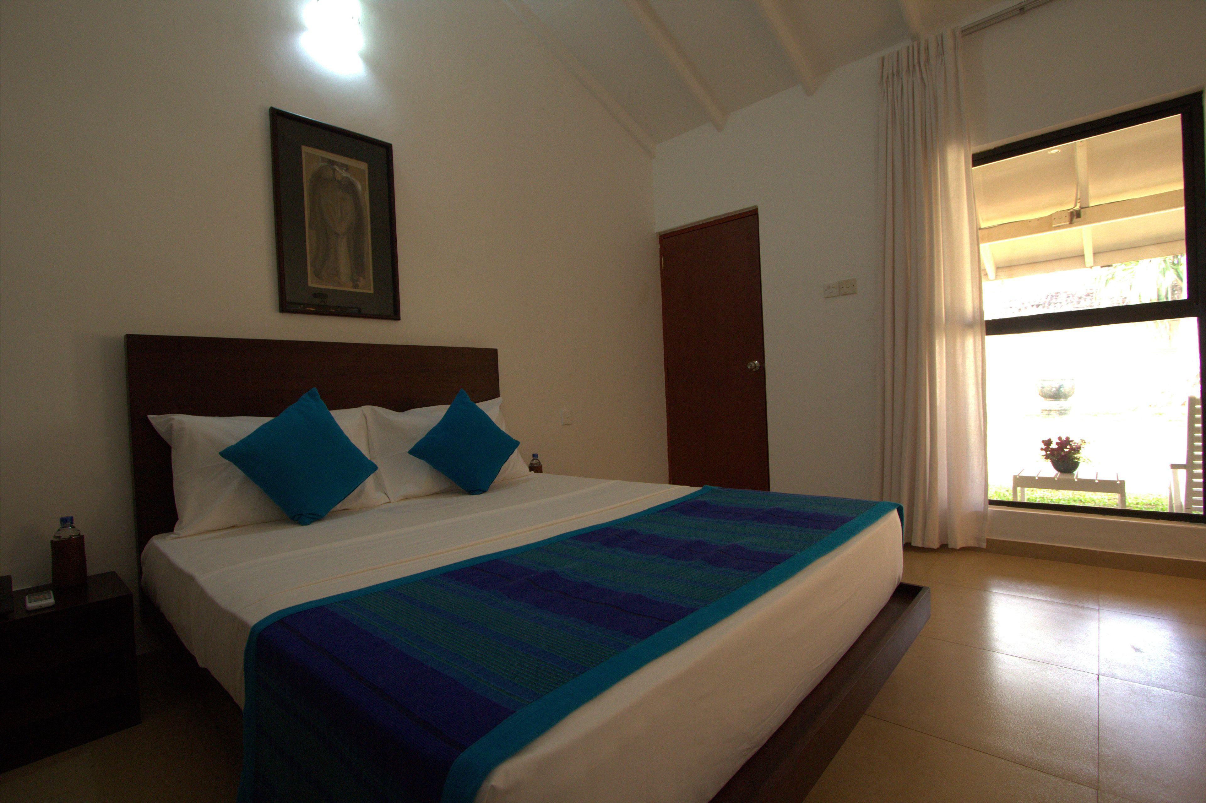 Quarto de hotel com uma cama grande, mesa de cabeceira, quadro na parede e janela com cortinas.