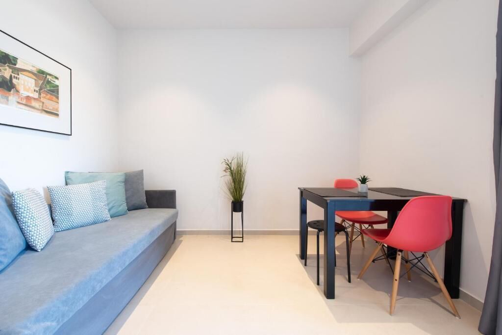 Quarto pequeno e moderno com um sofá azul, uma mesinha com cadeiras vermelhas e um vaso de planta.