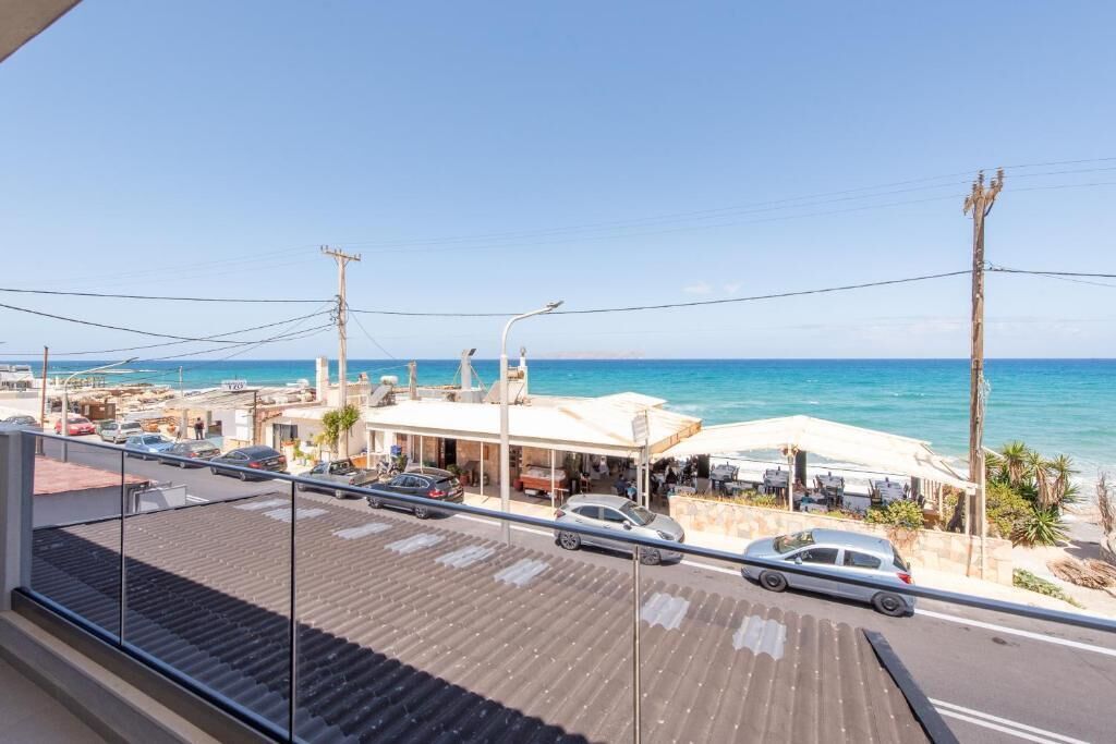 A coastal view from a balcony overlooking a street with parked cars, a beachside restaurant, and a clear blue sky.