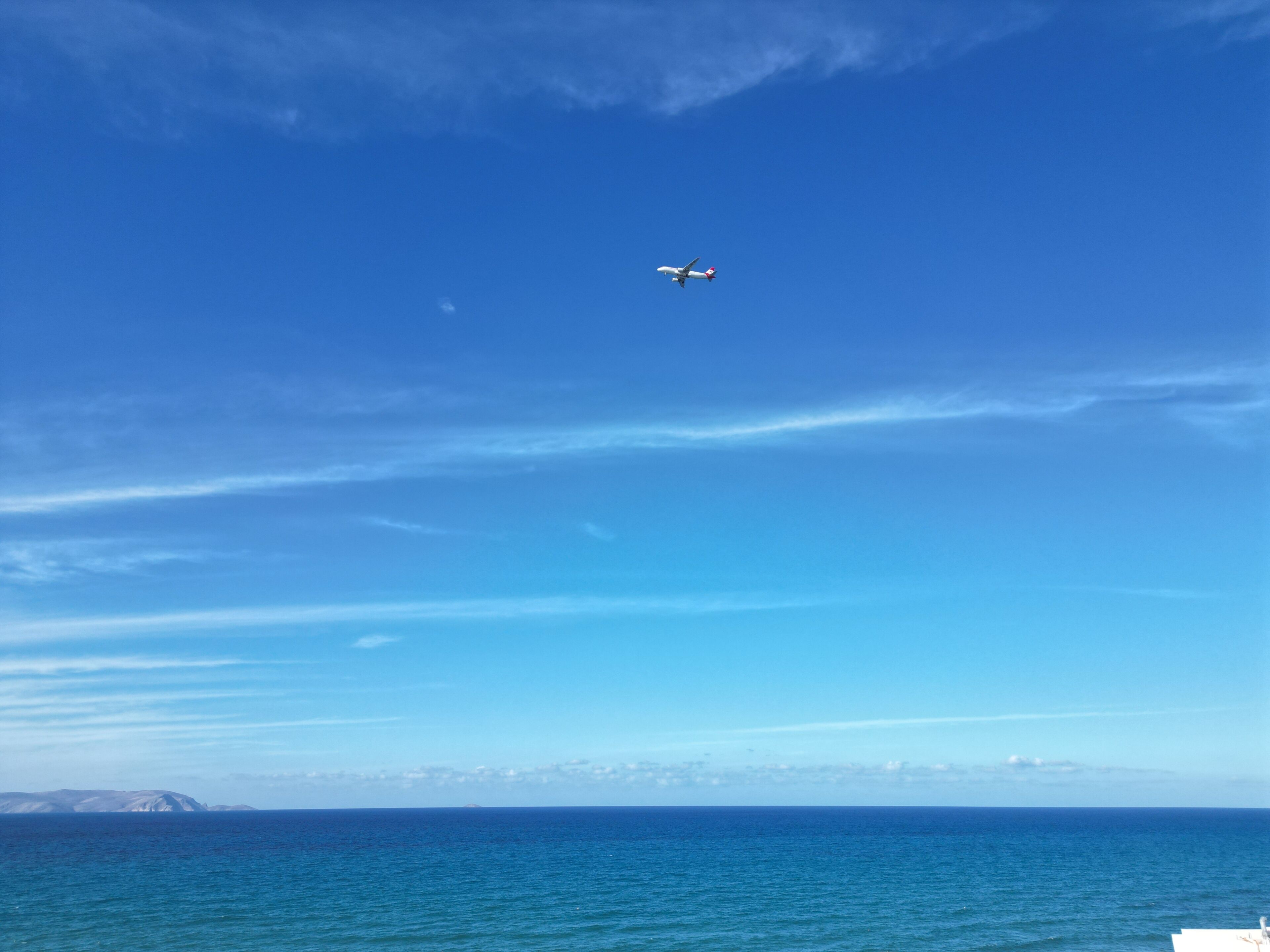 A clear blue sky with a plane flying over a calm sea.