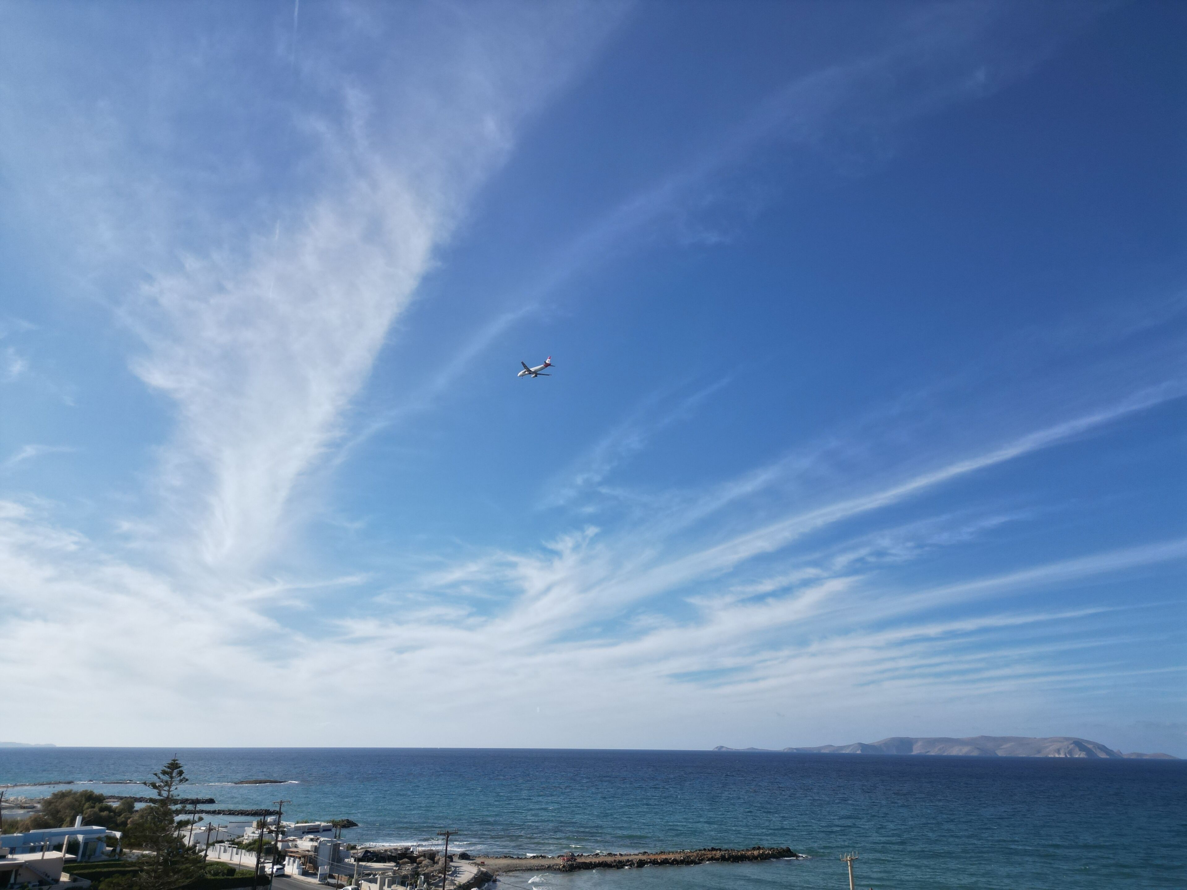 Vista costera con cielo despejado, un avión comercial en vuelo y una masa de tierra a lo lejos.
