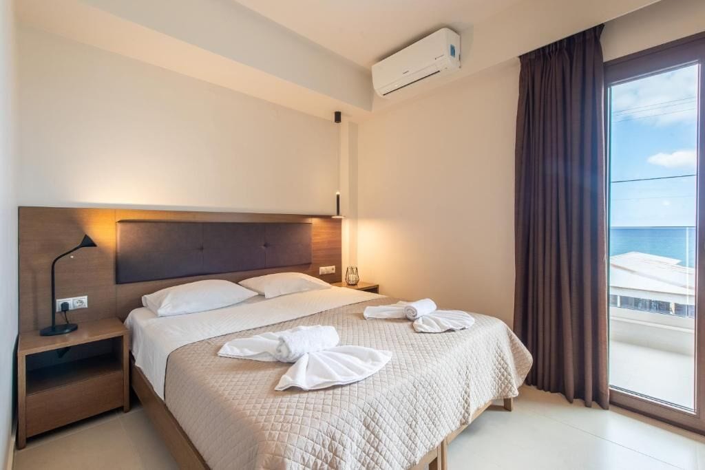 A hotel room with a bed, a nightstand, an air conditioning unit, a window with curtains, and a view of the sea.