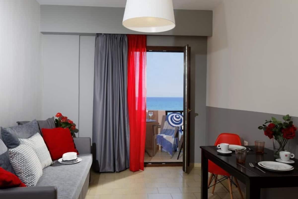Un salon moderne avec un canapé gris, des rideaux rouges et gris, une table à manger avec de l’assiette et des tasses blanches, et une vue sur la mer par une porte vitrée.