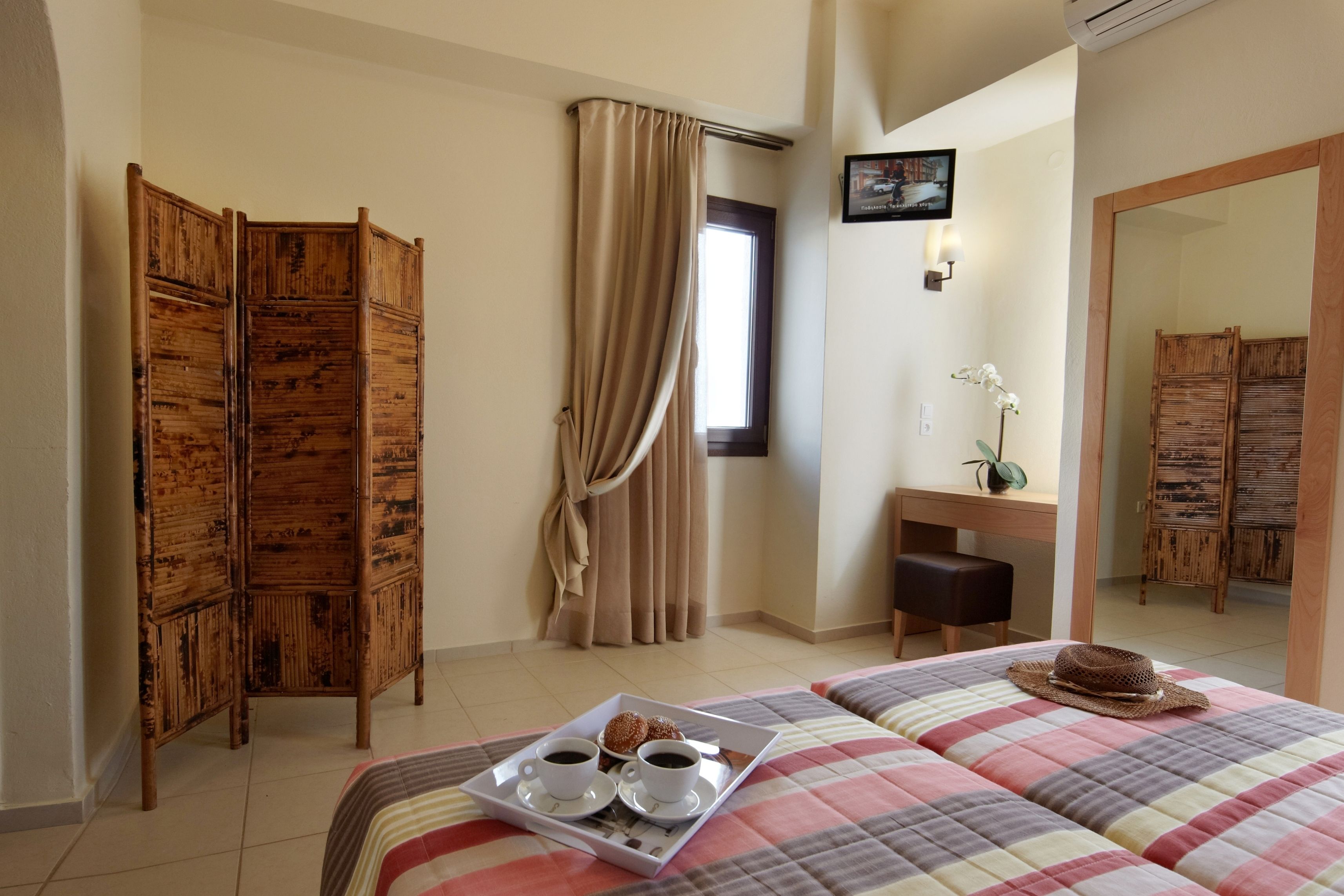 A hotel room with a bed, a wooden wardrobe, a desk with a chair, a mirror, and a window with curtains.