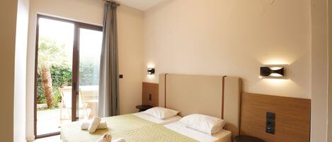 A hotel room with a bed, a nightstand, a lamp, a sliding glass door to the balcony, and a view of greenery outside.