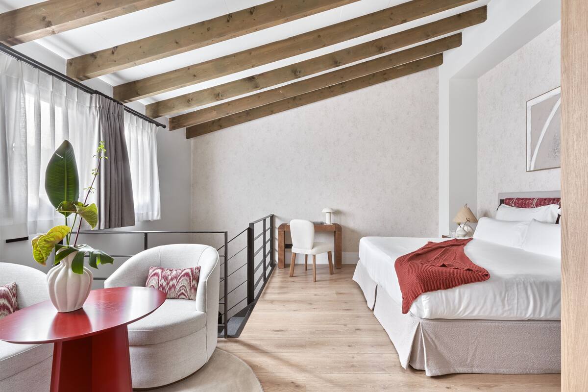 A modern, well-lit room with a wooden ceiling, a bed with white bedding, a red table, a white armchair, and a large plant.