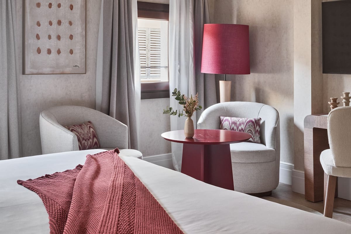 A hotel room with a large bed, two armchairs, a small table with a vase, a red lamp, and a painting on the wall.