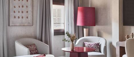 Habitación de hotel con una cama grande, dos sillones, una mesita con un florero, una lámpara roja y un cuadro en la pared.