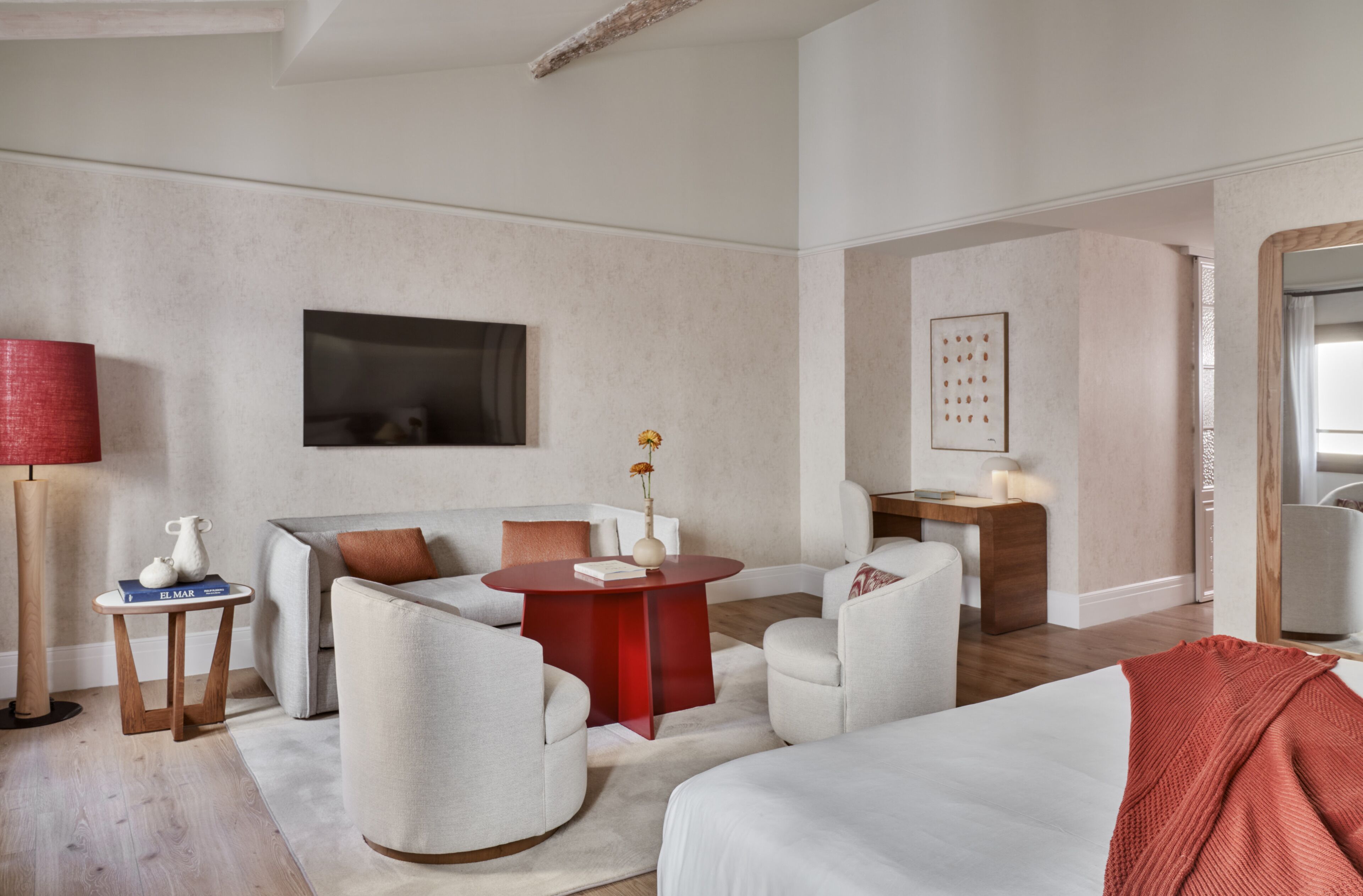 Una habitación de hotel moderna con sofá, sillones, una mesa redonda de madera y una cama con una manta roja.