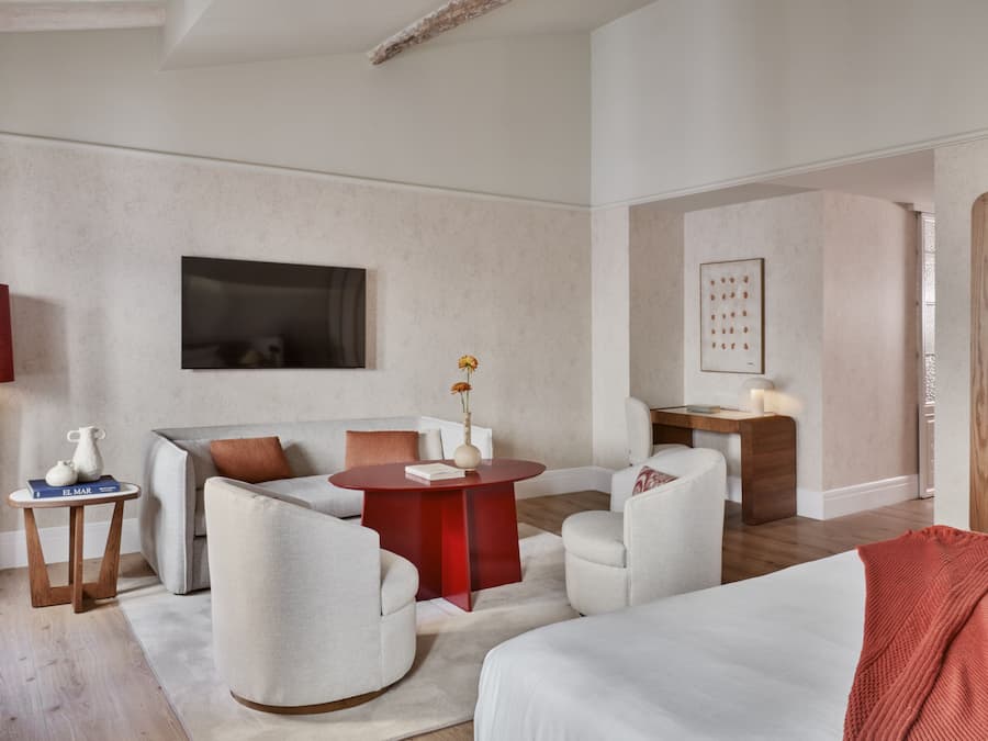 Una habitación de hotel moderna con sofá, sillones, una mesa redonda de madera y una cama con una manta roja.