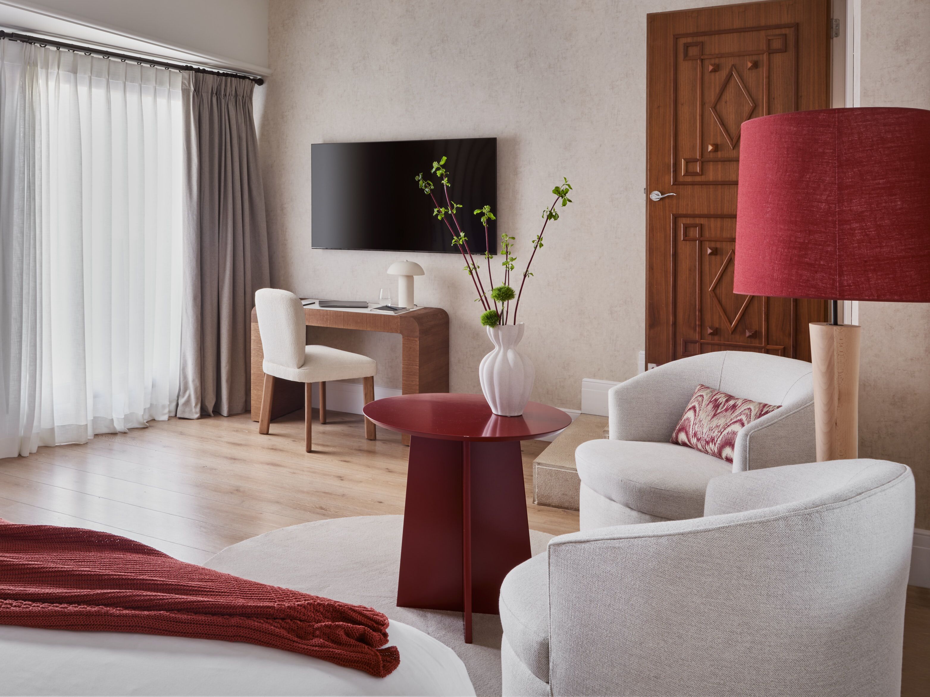 Una habitación de hotel moderna con un escritorio de madera, un televisor, una lámpara roja y un sillón blanco.