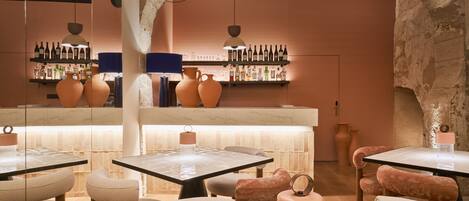 Interior de restaurante con mesas de mármol, sillas acolchadas y una barra de vinos con estantes llenos de botellas.