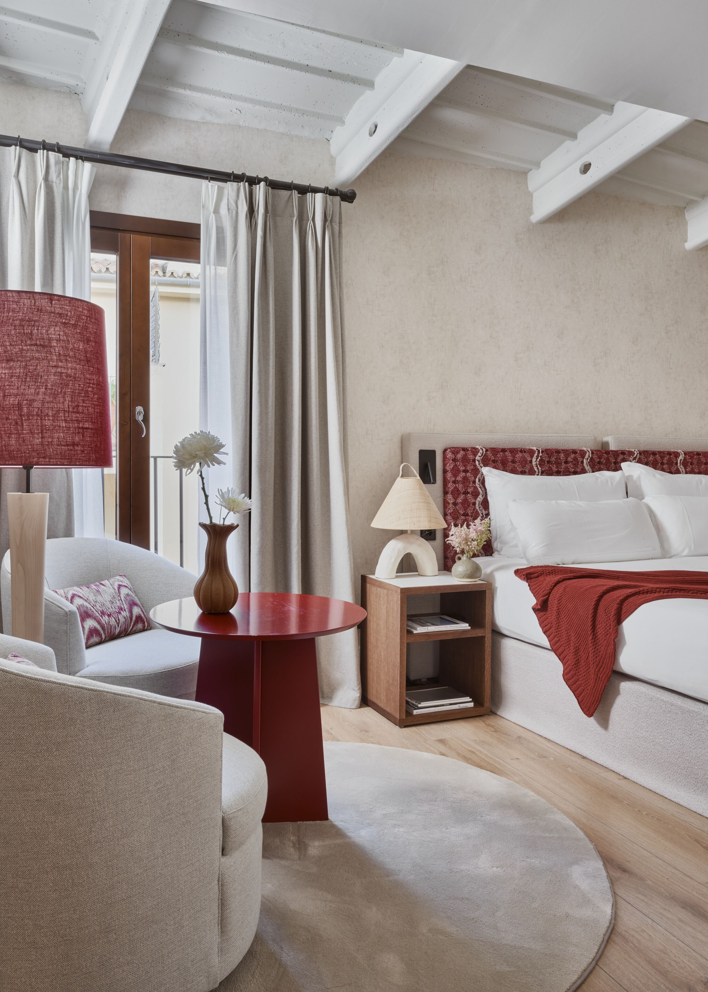 A hotel room with a bed, a sitting area with a chair and a small table, a nightstand with a lamp, and a view of the outside through a window with curtains.