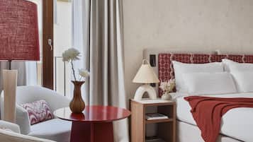 A hotel room with a bed, a sitting area with a chair and a small table, a nightstand with a lamp, and a view of the outside through a window with curtains.