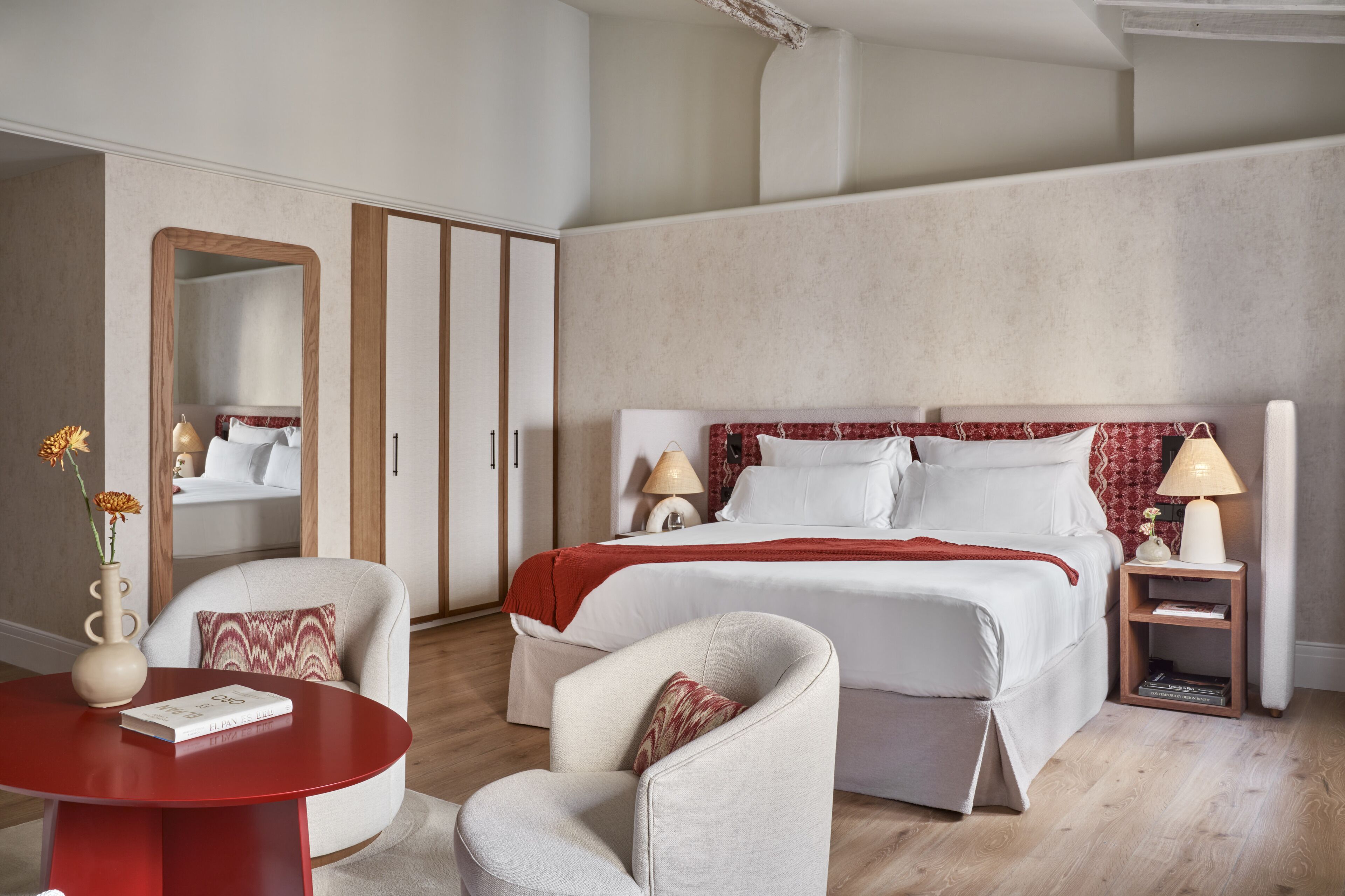 Quarto de hotel com uma cama, duas cadeiras, uma mesinha, um espelho e uma mesinha de cabeceira com luminárias.