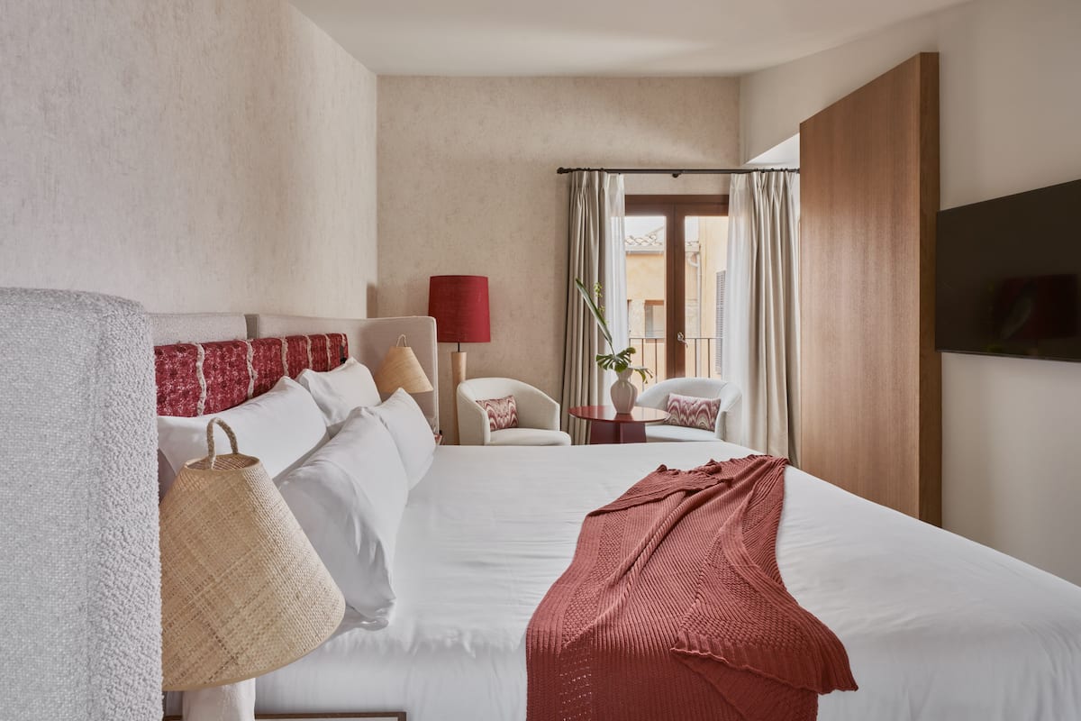 A hotel room with a large bed, a sitting area with a chair and a table, a TV mounted on the wall, and a view through a window with curtains.
