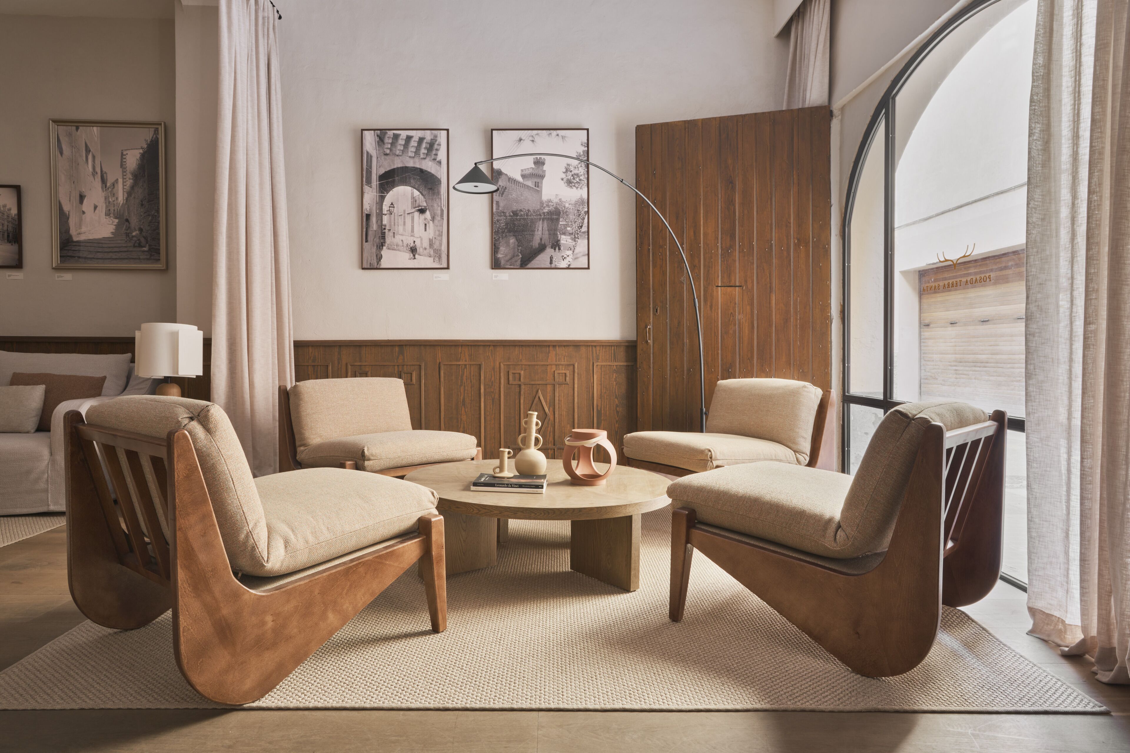 Una sala de estar moderna con una mesa redonda, dos sillones y un sofá. Hay cuadros enmarcados en la pared, una lámpara y vistas al exterior a través de un gran ventanal.