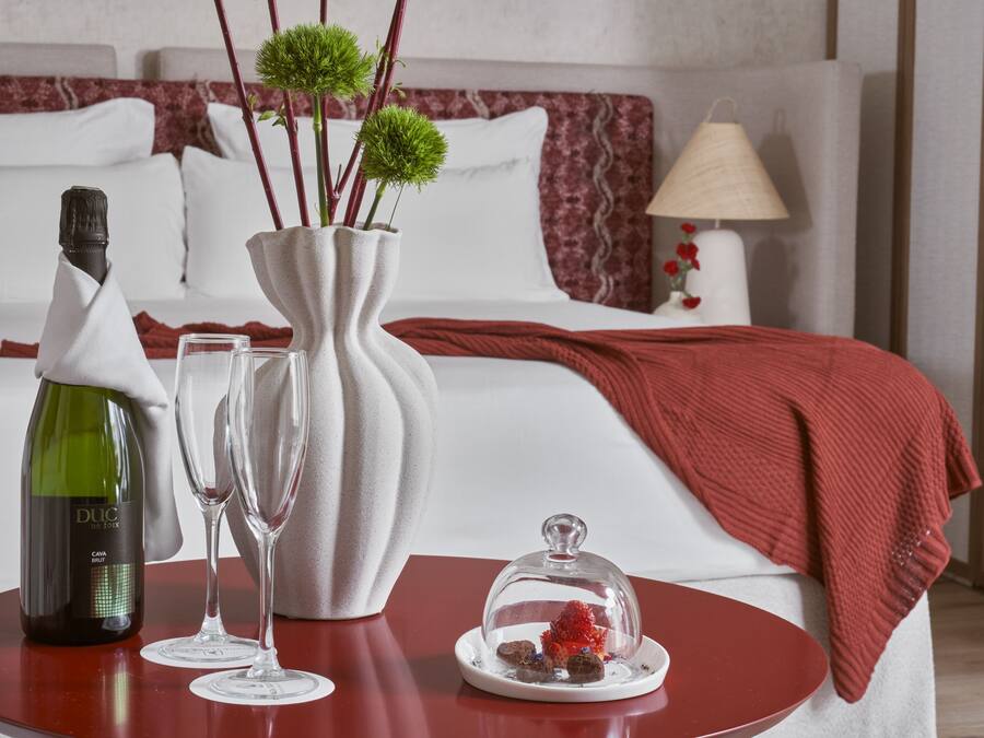 A hotel room with a bed, a bedside table with a bottle of wine, two glasses, a vase with green branches, and a decorative item on the table.