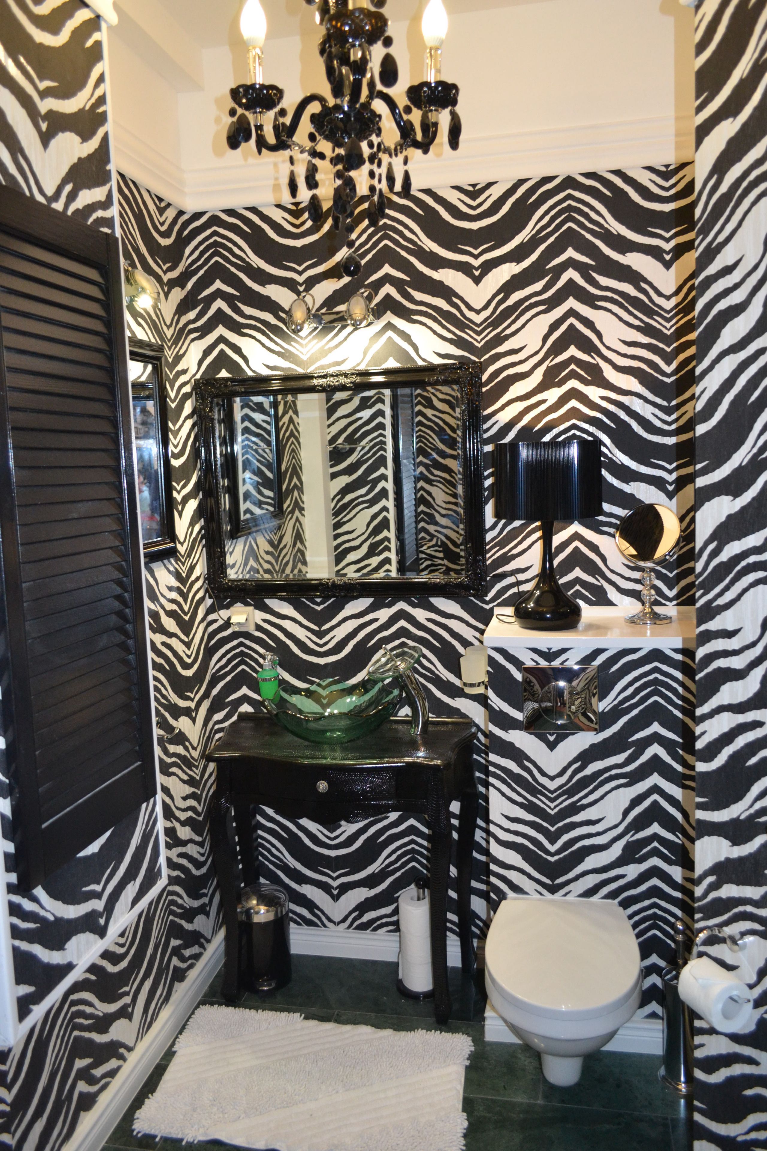 Une salle de bain avec un papier peint à rayures zébrées, un lustre, un miroir, un lavabo avec un récipient en verre vert, des toilettes et une petite table avec une lampe.