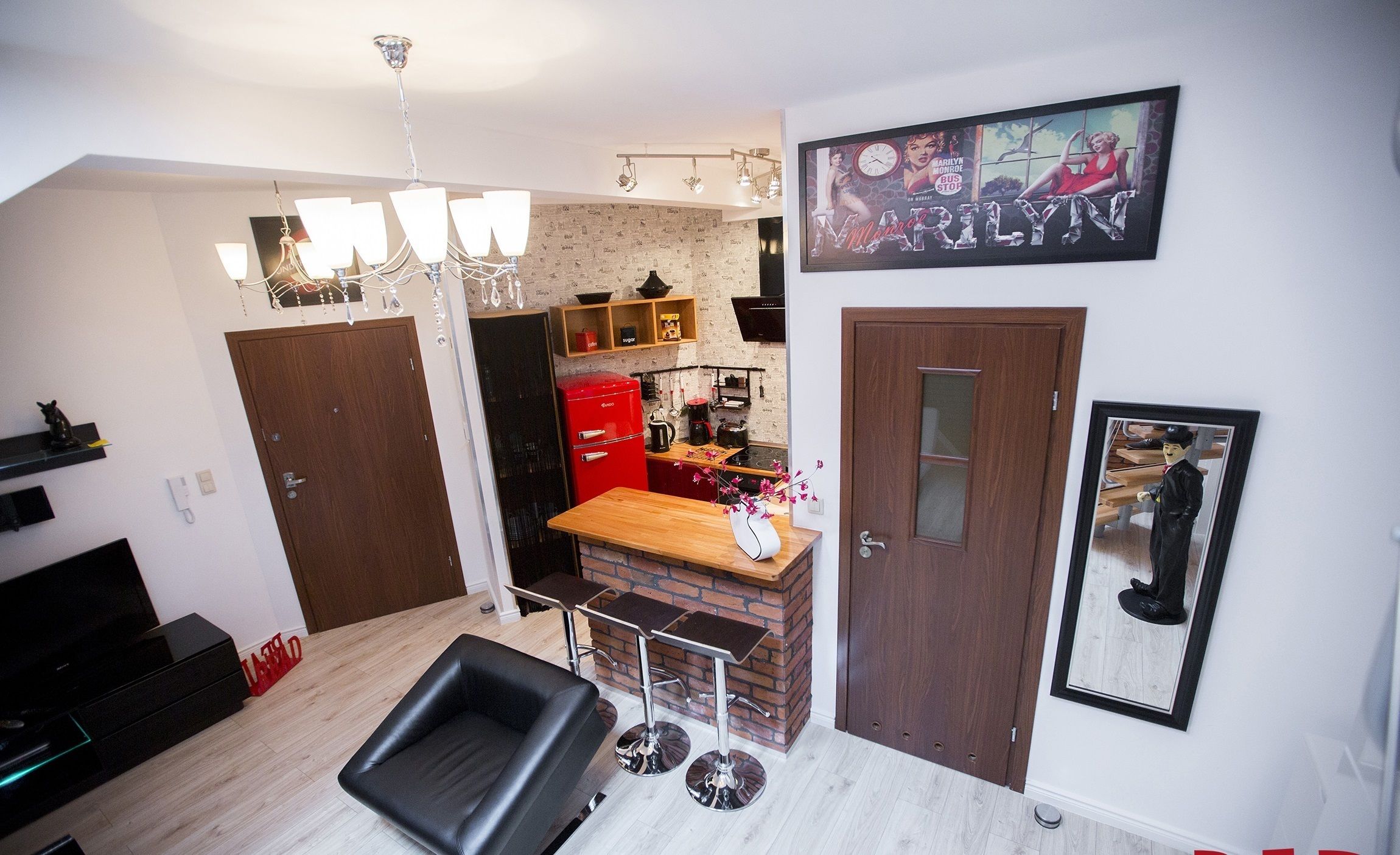 Una cucina moderna con zoccolo in mattoni, un frigorifero rosso e un piano di lavoro in legno. Nell'area living, un divano in pelle nera, un lampadario e un quadro di Marilyn Monroe appeso al muro.