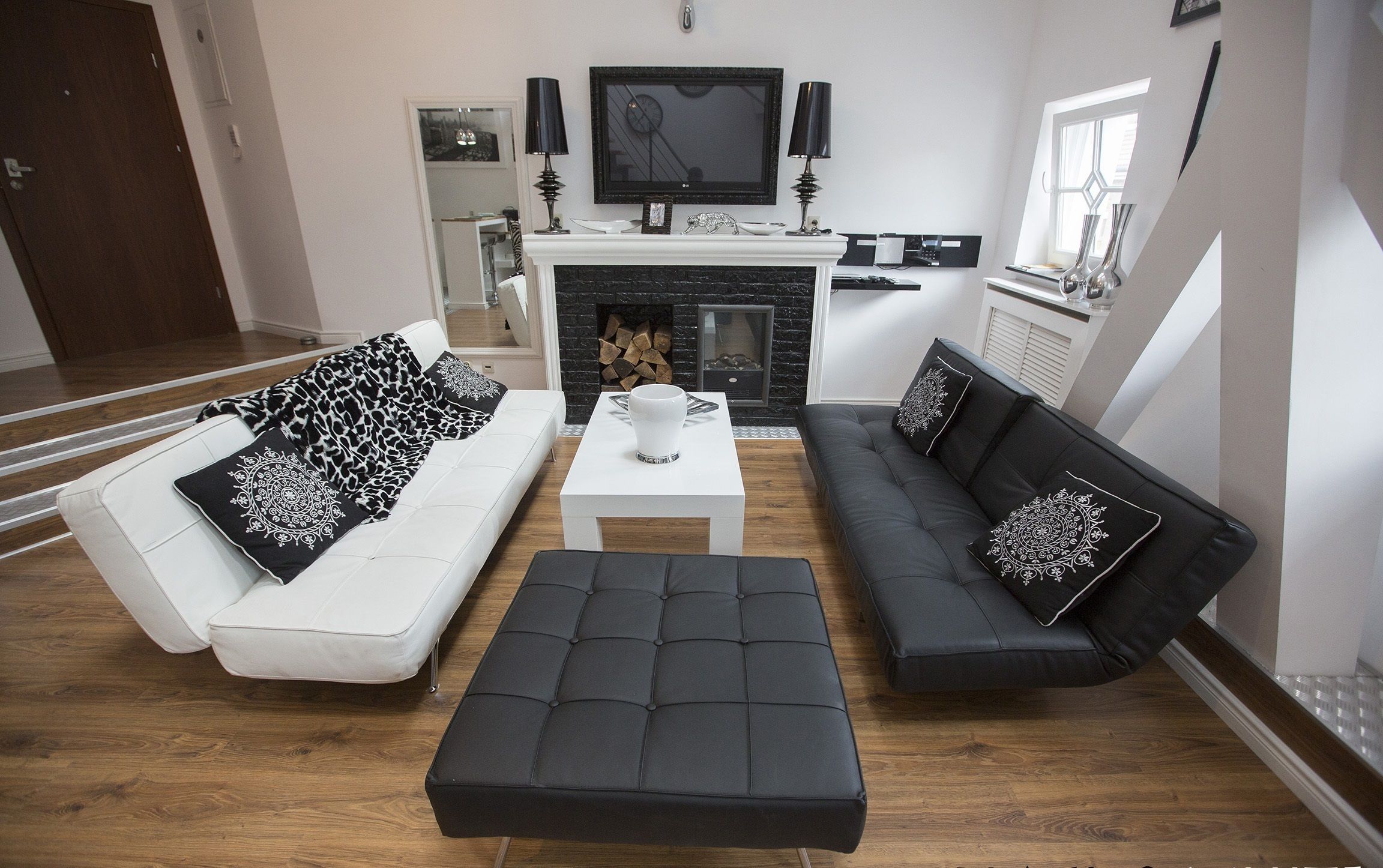 Una sala de estar moderna con un sofá blanco, un sofá negro y un reposapiés negro. Hay una chimenea con un televisor encima, y un comedor con una mesa y sillas.