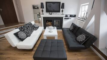 Una sala de estar moderna con un sofá blanco, un sofá negro y un reposapiés negro. Hay una chimenea con un televisor encima, y un comedor con una mesa y sillas.