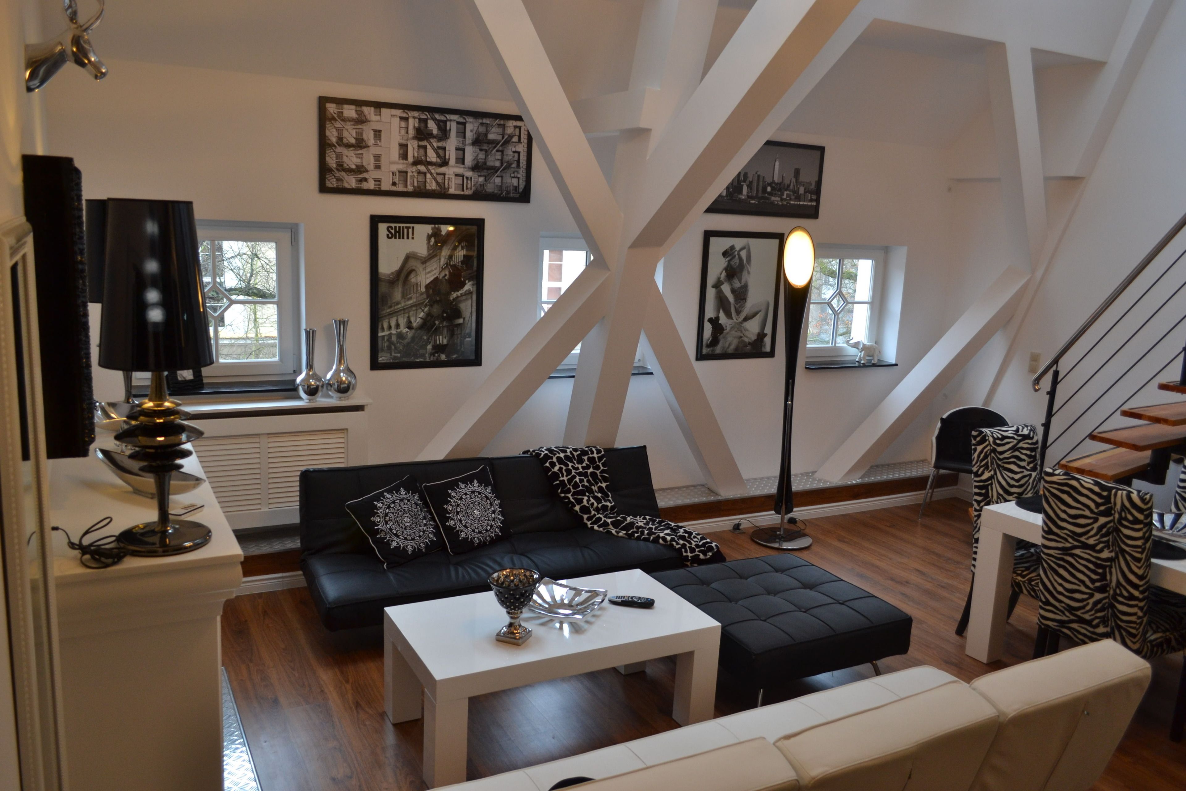 A modern loft-style living room with a black leather sofa, white coffee table, and zebra-patterned chairs. There are framed artworks on the walls and a staircase leading to an upper level.