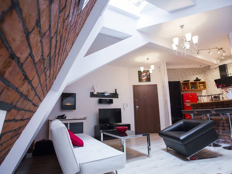 A modern loft apartment with a sloped ceiling, a dining area with a red table and chairs, a kitchen with a brick backsplash, and a living room with a white sofa and a black chair.