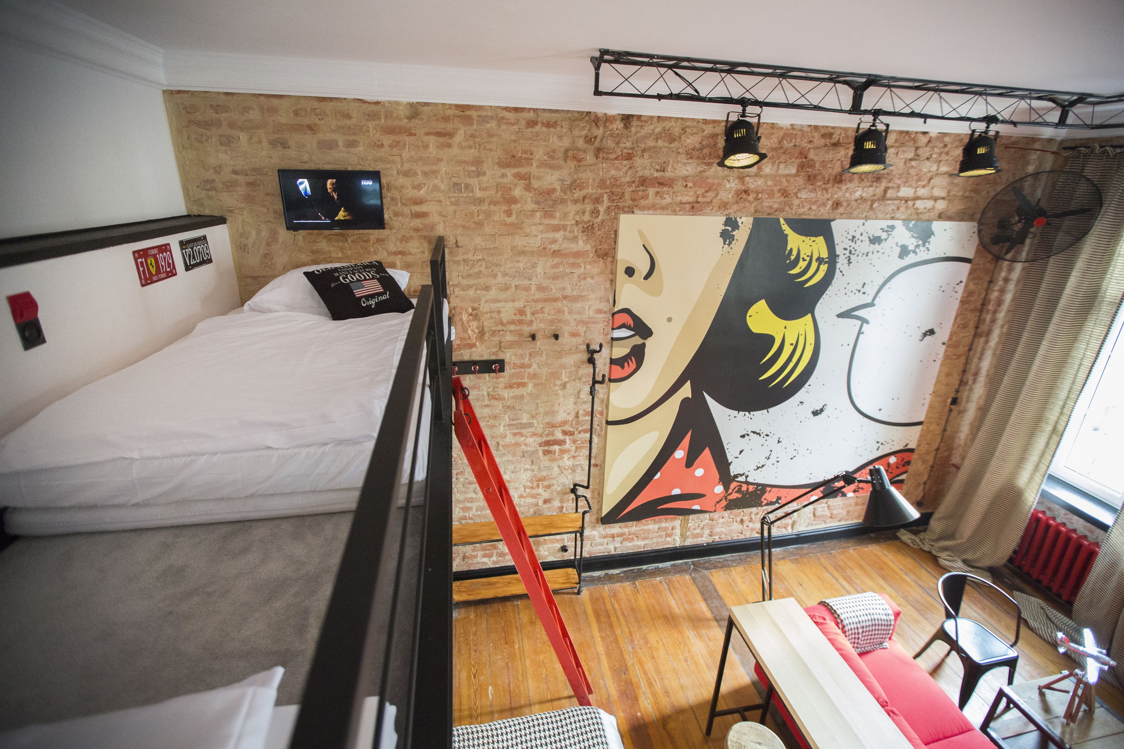A room with a bed, a red ladder, a table with a red cushion, a black chair, and a large abstract painting on the wall.