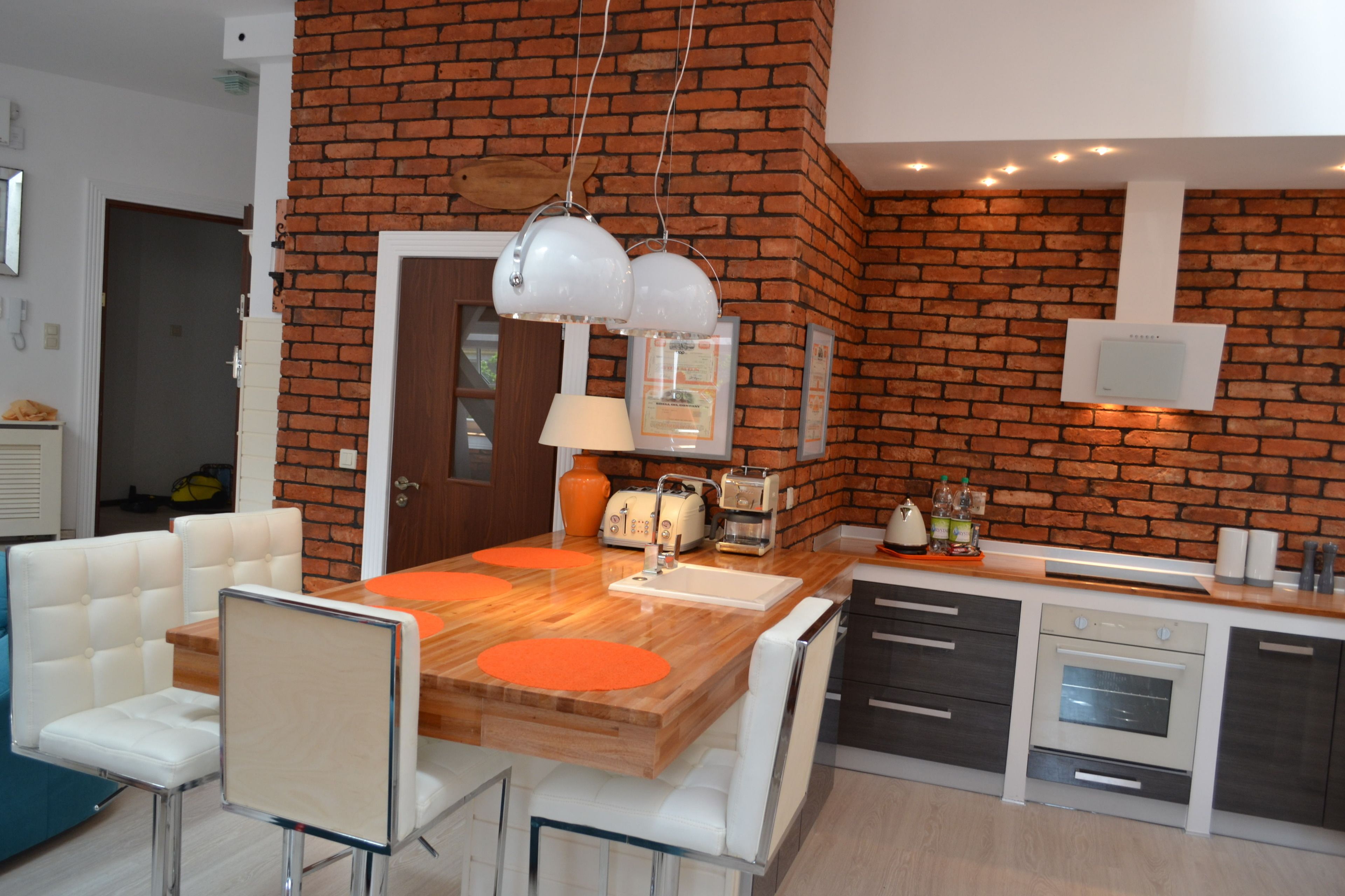Eine moderne Küche mit Backsteinwand, weißen Schränken und einer Holztischinsel mit orangefarbenen Untersetzern.