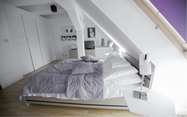 Une chambre moderne avec un plafond de style industriel, un lit recouvert d’une couette grise et blanche, une table de chevet avec une lampe et un téléviseur posé sur un meuble.