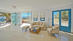 A spacious living room with a large glass door leading to an outdoor area, a beige sofa, a striped armchair, and a coffee table. - Dilino Hotel (Naxos)