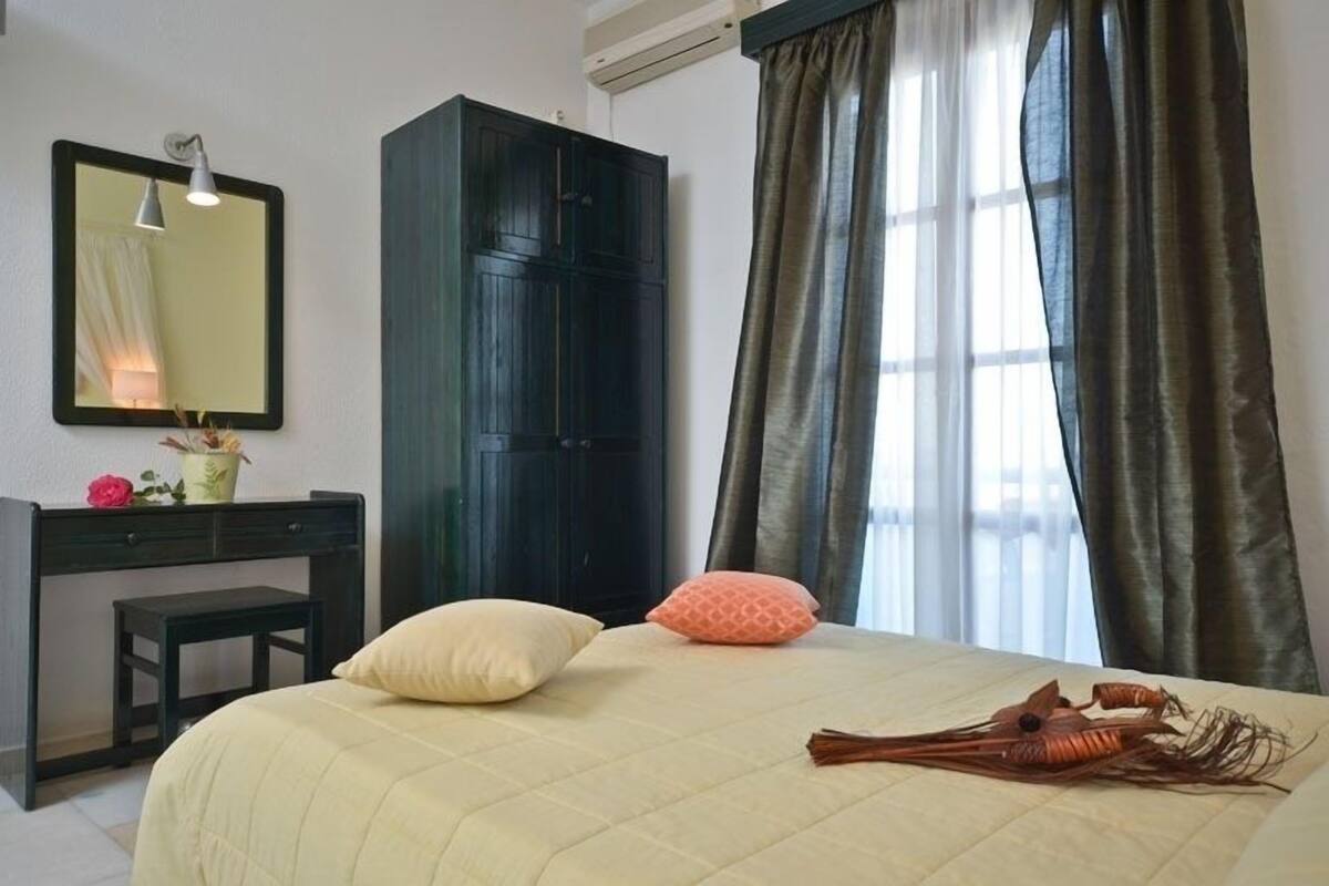 A bedroom with a bed, a dark wardrobe, a desk with a chair, a mirror, and a window with curtains.
