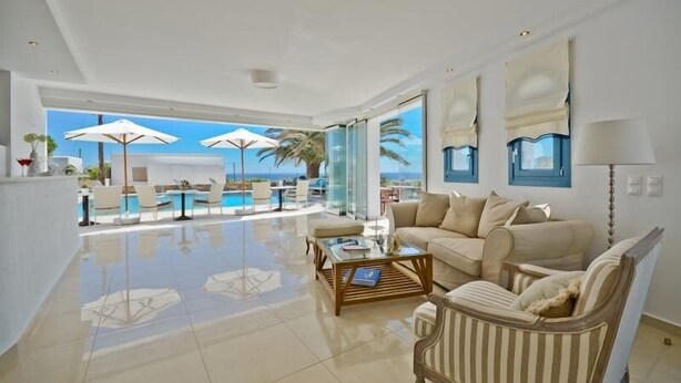 A spacious living room with a large window, a sofa, a coffee table, and a view of the pool and ocean. - Dilino Hotel (Naxos)