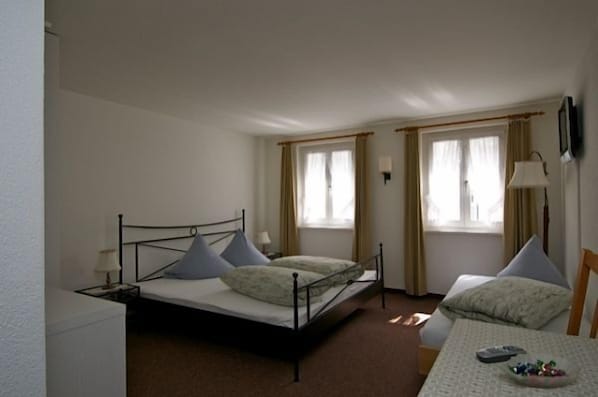 A single bed with a metal frame, two bedside tables, a small table with a bowl of snacks, and two windows with curtains. - Gasthaus Skiklub (Andermatt)
