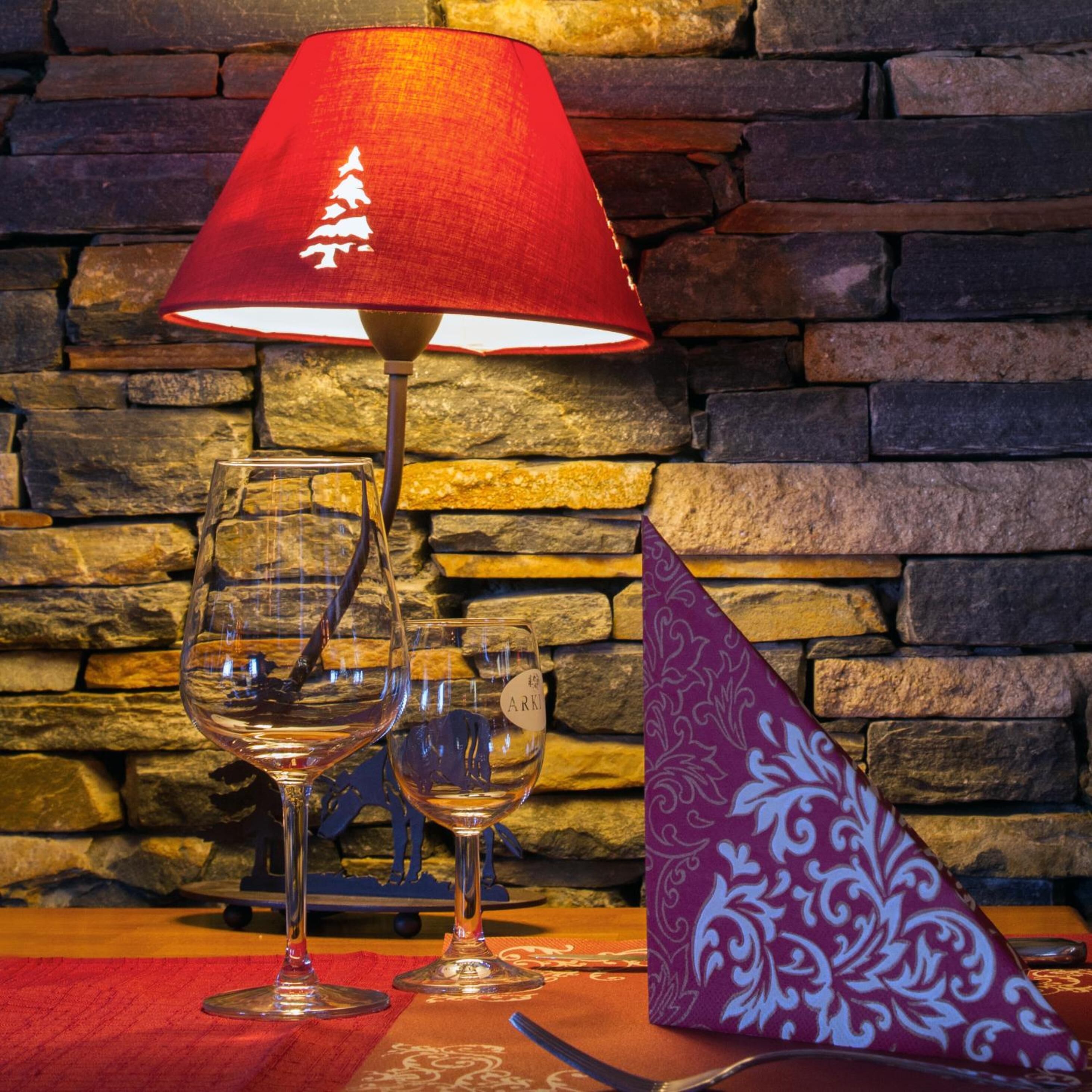 A table with wine glasses and a napkin, a red lamp, and a stone wall background.